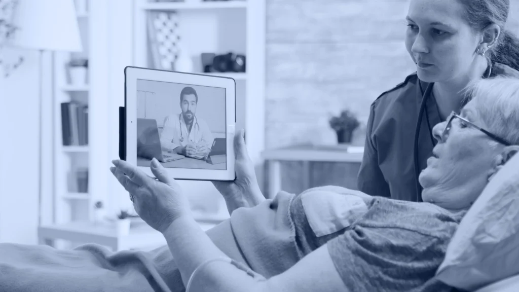 Paciente adulta mayor en cama viendo una teleconsulta en tablet junto a su cuidadora