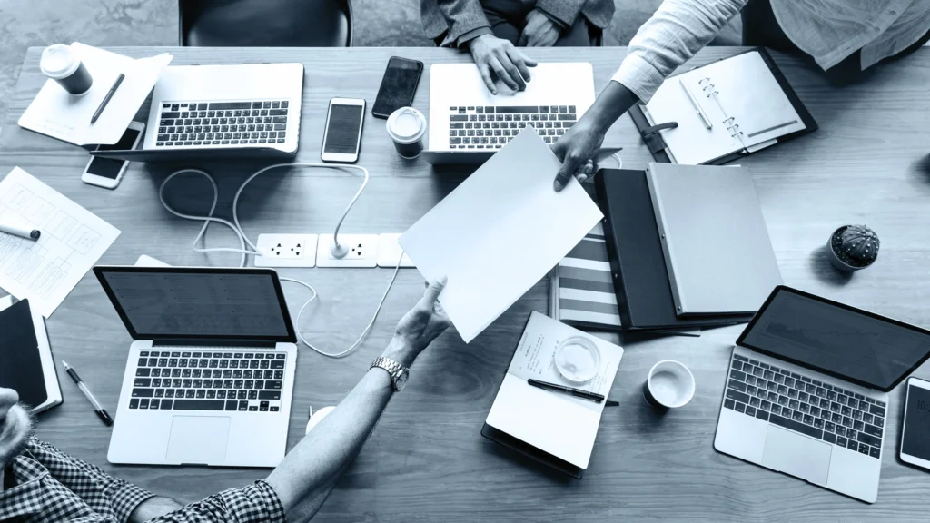 Equipo de trabajo intercambiando documentos sobre una mesa con notebooks